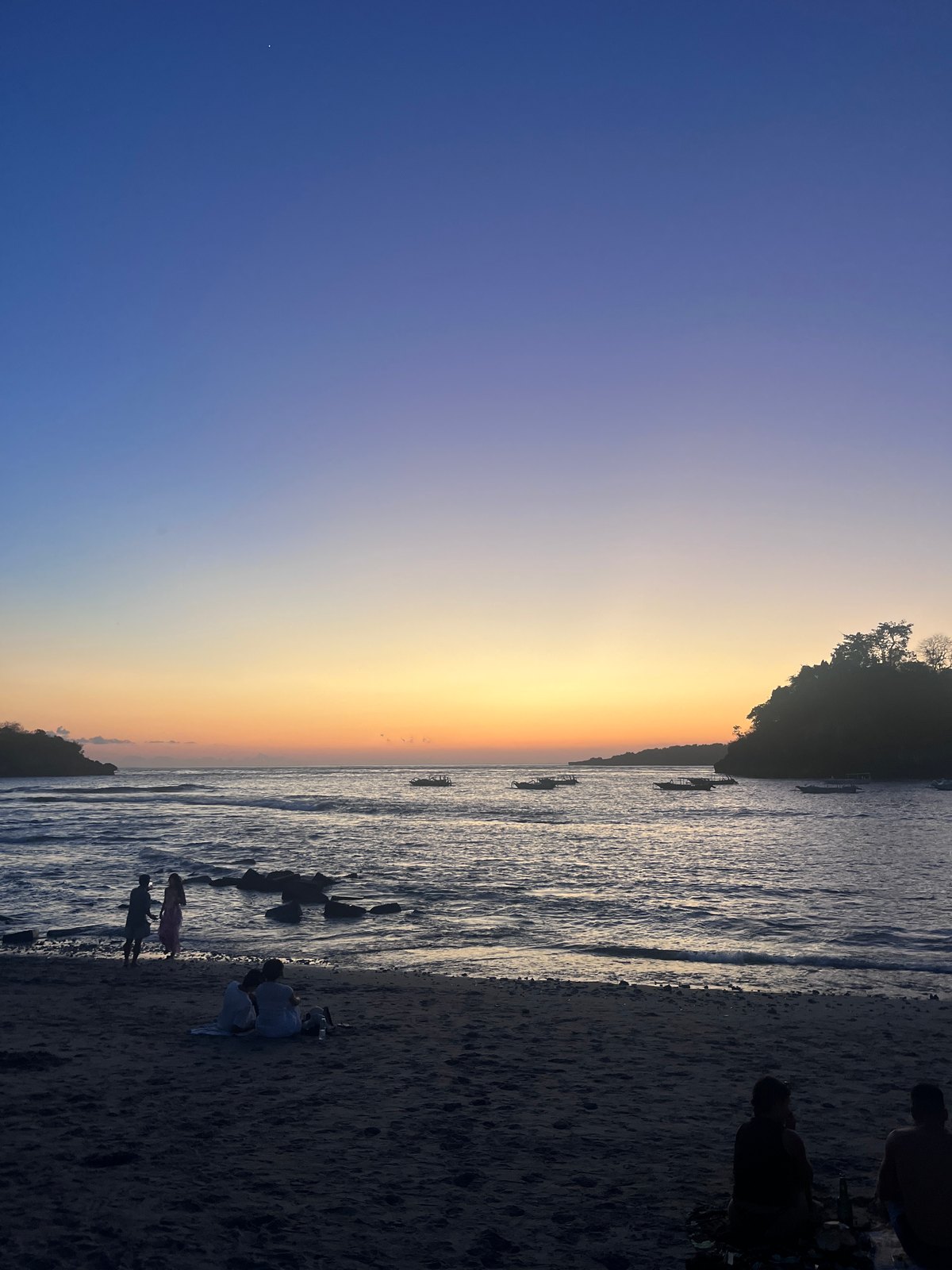 People sitting on Crystal Bay beach with boats offshore at dusk.