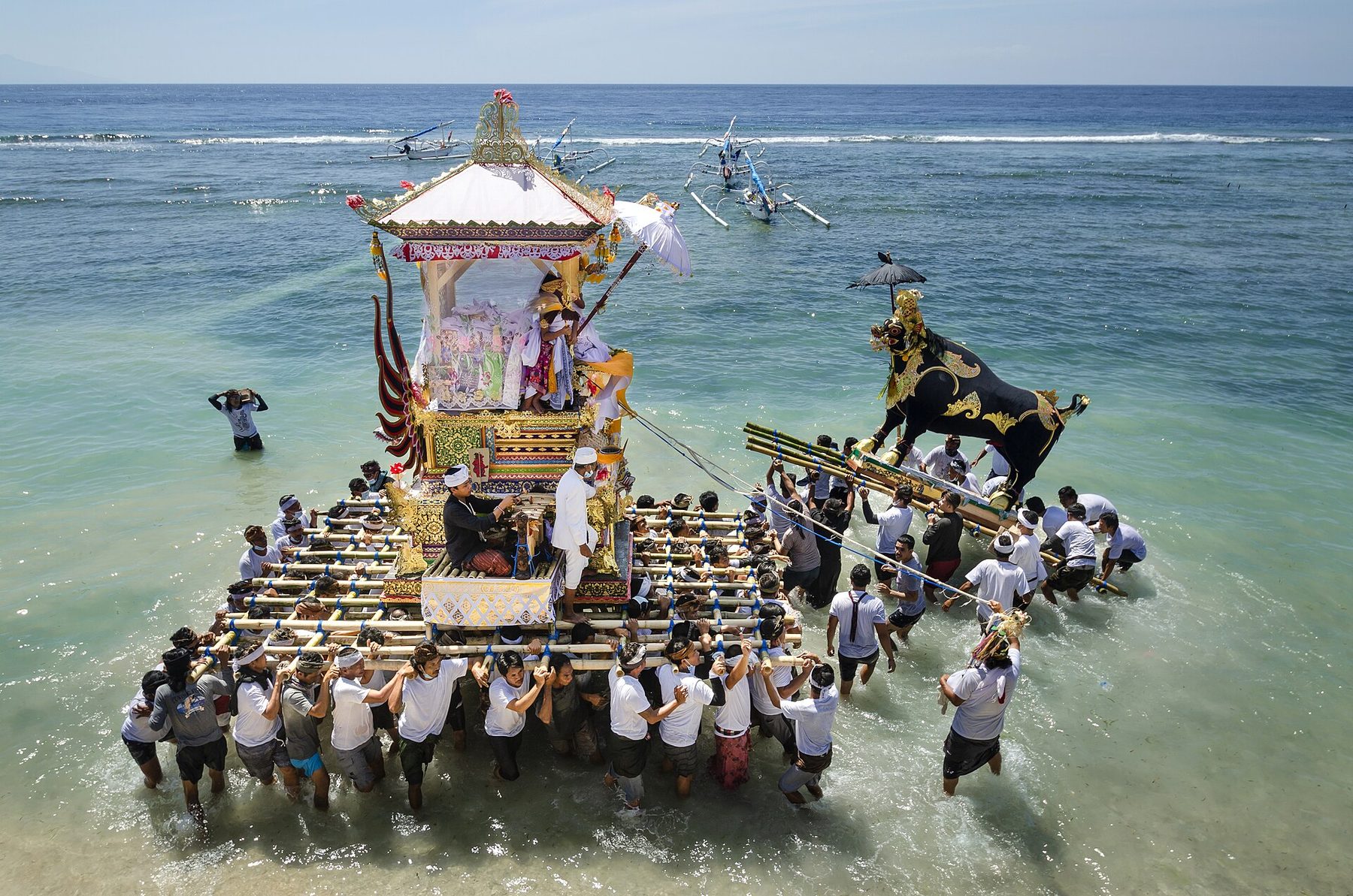 Men in white carry a tall cremation tower and a black bull figure on a bamboo platform through shallow sea water during a ngaben in Nusa Penida.