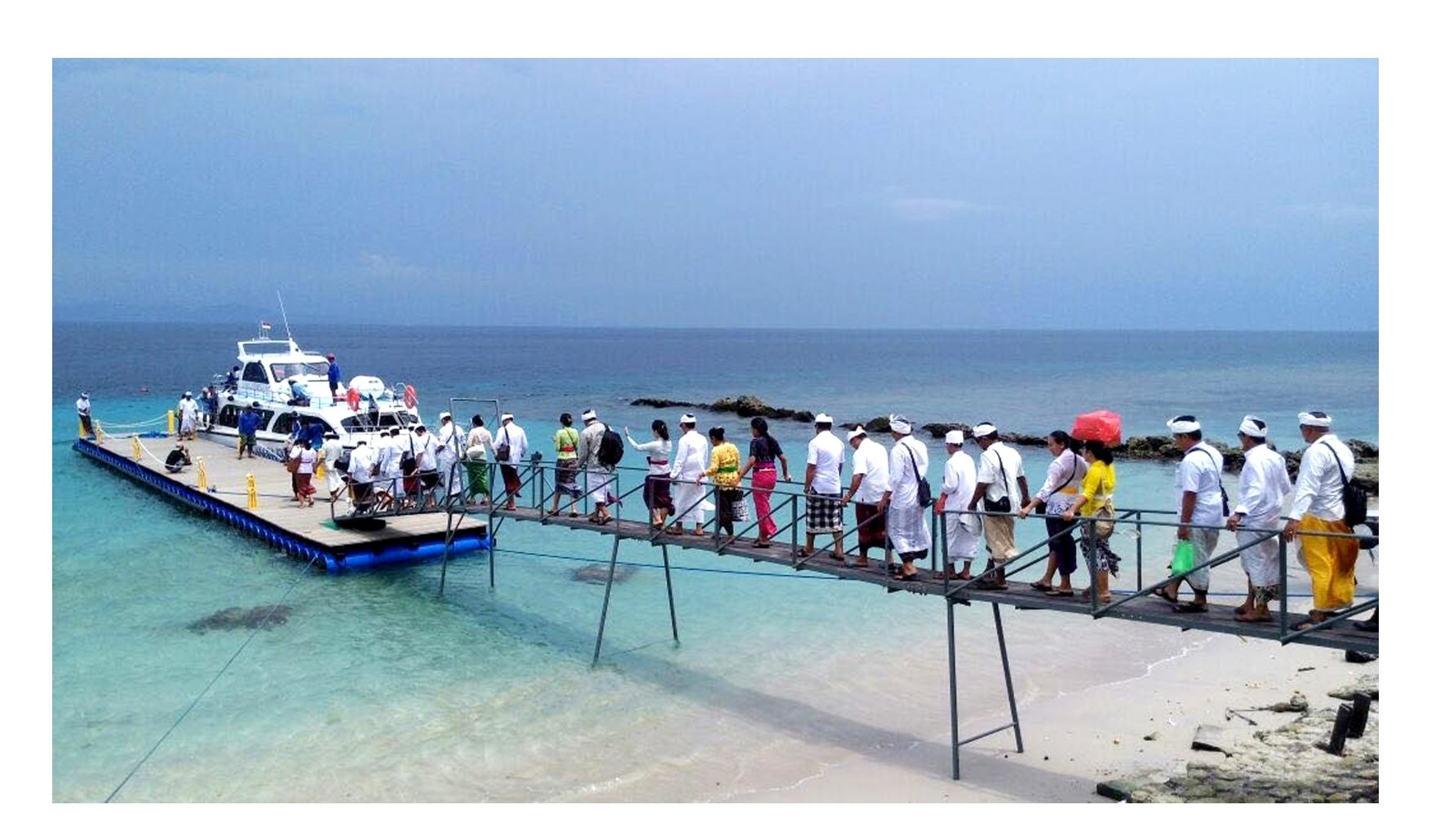 People in white Balinese temple dress walk along a footbridge onto a floating pier at Sampalan, with a speedboat moored at the end.