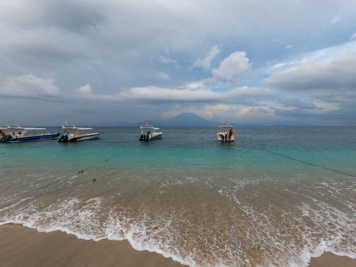 Small boats moored off a sandy beach on Nusa Penida's north coast, with Mount Agung on Bali under cloud across the strait.
