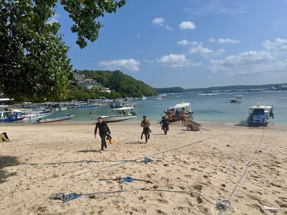Divers in wetsuits walking up the sand at Toyapakeh beach from a moored dive boat.