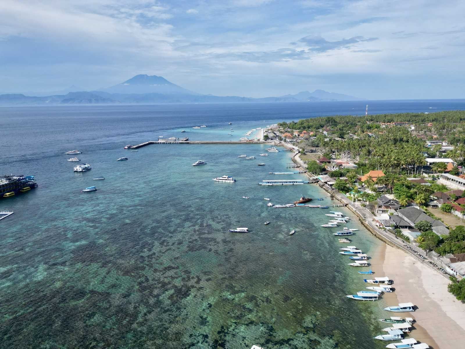 Aerial view of Toyapakeh's beach lined with small boats, the long harbour jetty beyond and Mount Agung on Bali across the strait.