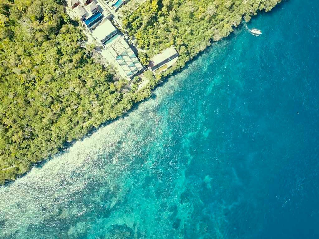 Aerial view of the Toyapakeh coast, with a pale shallow reef along the shore dropping into deep blue water and a boat offshore.