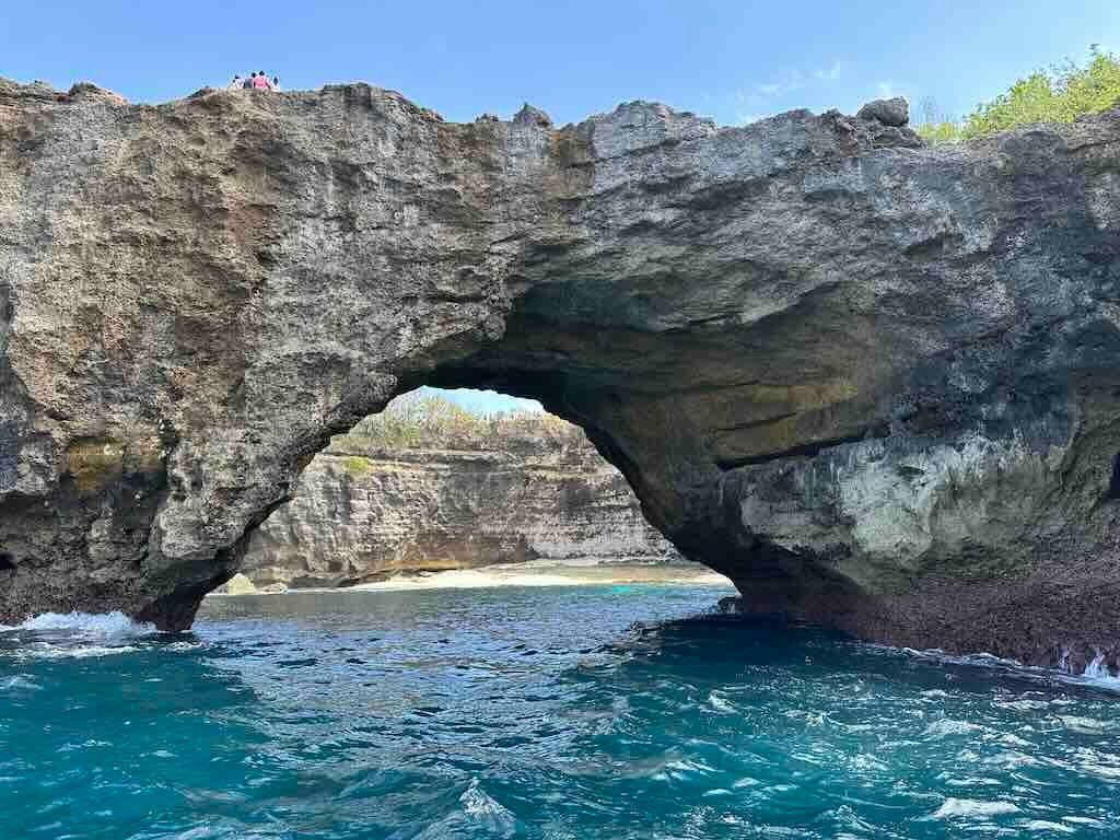 The rock arch of Broken Beach seen from the water, with the sandy cove visible through the opening and people standing on top.