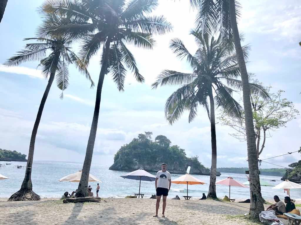 Palm trees, umbrellas and visitors on the sand at Crystal Bay, with the wooded rock island offshore.