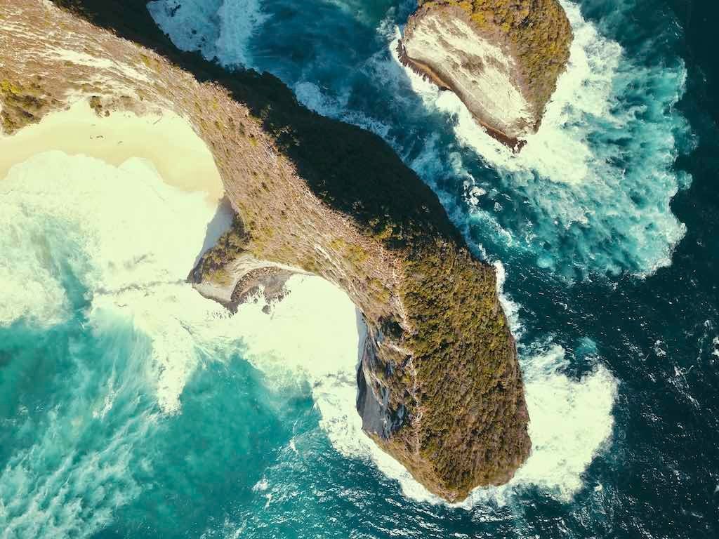 Drone view straight down onto Kelingking's long limestone headland, with waves breaking on the beach and the rocks below.